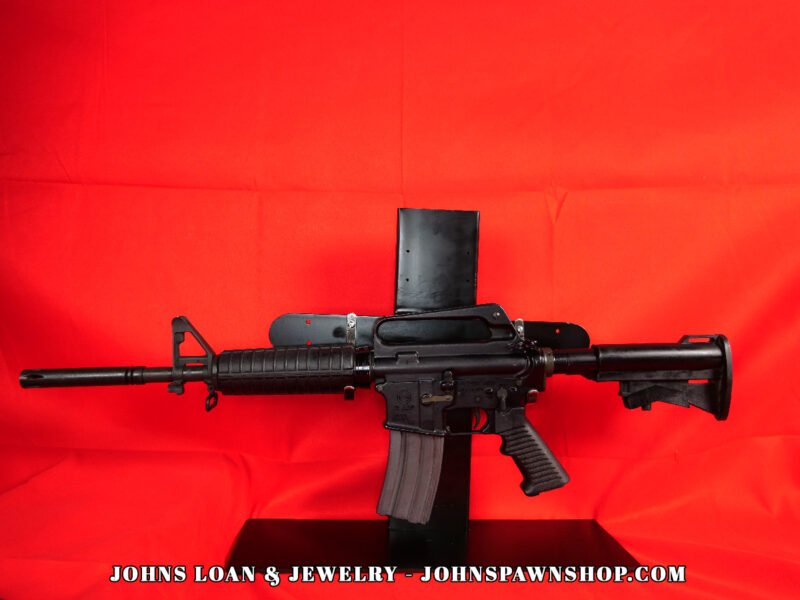 Olympic Arms CAR-15 semi-auto rifle, a vintage-style firearm, displayed against a red backdrop at John's Loan & Jewelry. The black AR-15 variant features a pinned flash hider and a 30+1 capacity magazine.