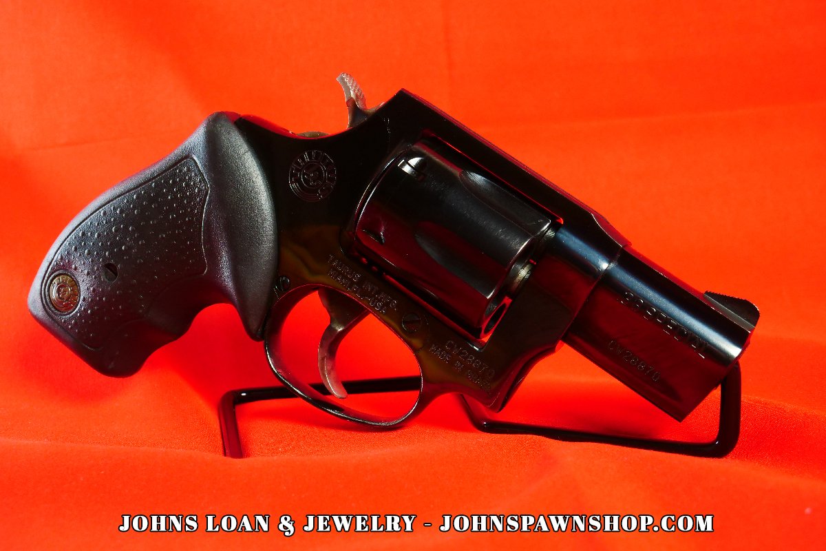 Side-profile view of a glossy black Taurus Model 85 snub nose revolver chambered in .38 Special, displayed on a red table by John's Loan & Jewelry.